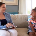 A therapist holds a clipboard during a session with a young child. They're both sitting on a couch and smiling at one another.