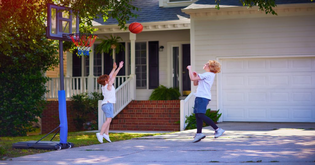 Two young boys, dressed in t-shirts and shorts, playing basketball in a driveway in front of a house.