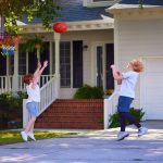 Two young boys, dressed in t-shirts and shorts, playing basketball in a driveway in front of a house.