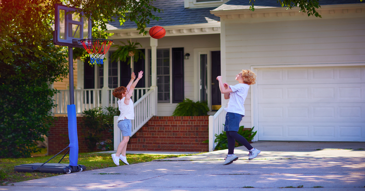 Two young boys, dressed in t-shirts and shorts, playing basketball in a driveway in front of a house.