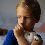 A blonde boy sucking thumb, holding a stuffed toy, with a blurred indoor background and soft lighting.