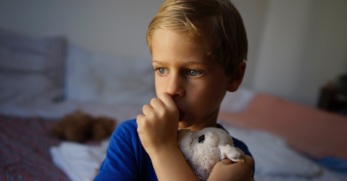 A blonde boy sucking thumb, holding a stuffed toy, with a blurred indoor background and soft lighting.