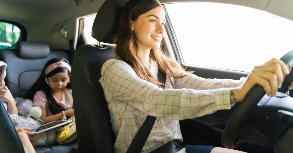 A woman smiles as she drives a vehicle with two children in the back. Both kids look at a storybook.