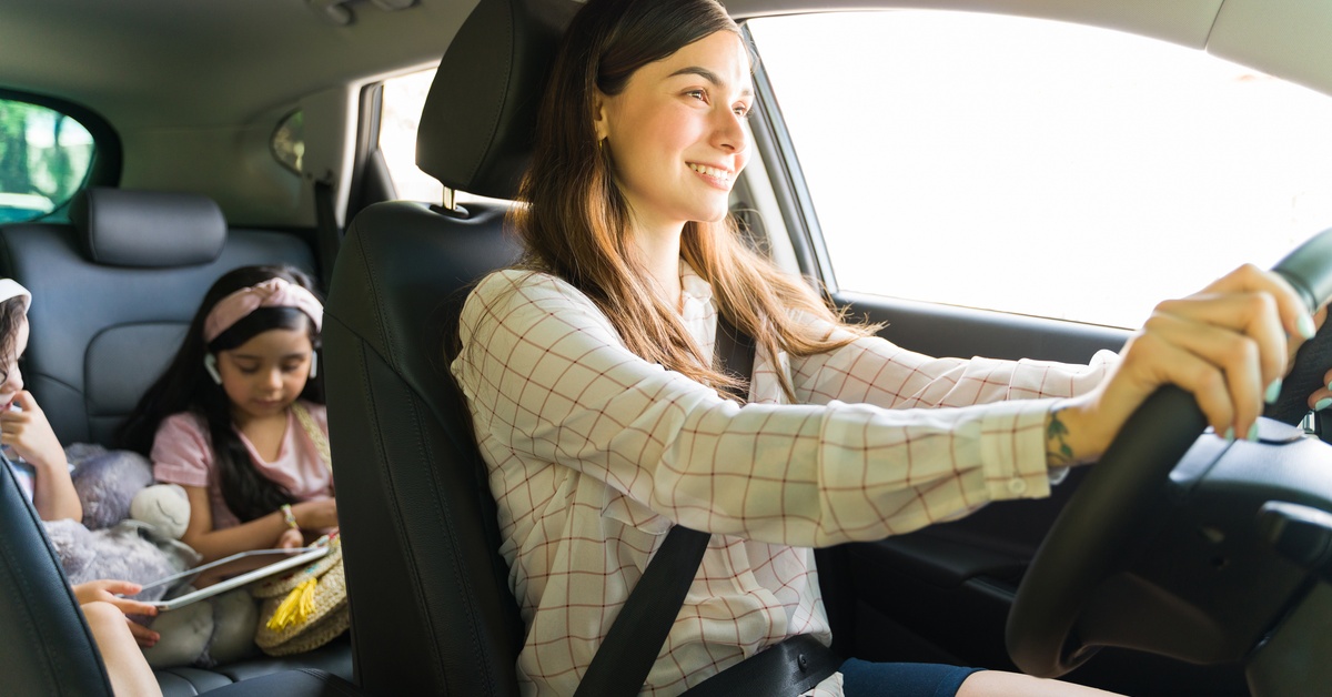 A woman smiles as she drives a vehicle with two children in the back. Both kids look at a storybook.