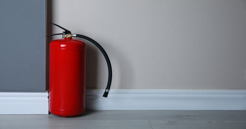 A red fire extinguisher on a light wooden floor, set against gray walls with a white baseboard in a minimalist room.
