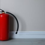 A red fire extinguisher on a light wooden floor, set against gray walls with a white baseboard in a minimalist room.