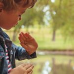 A little boy standing outdoors near a lake, holding a ladybug on the side of his palm and looking down at it.
