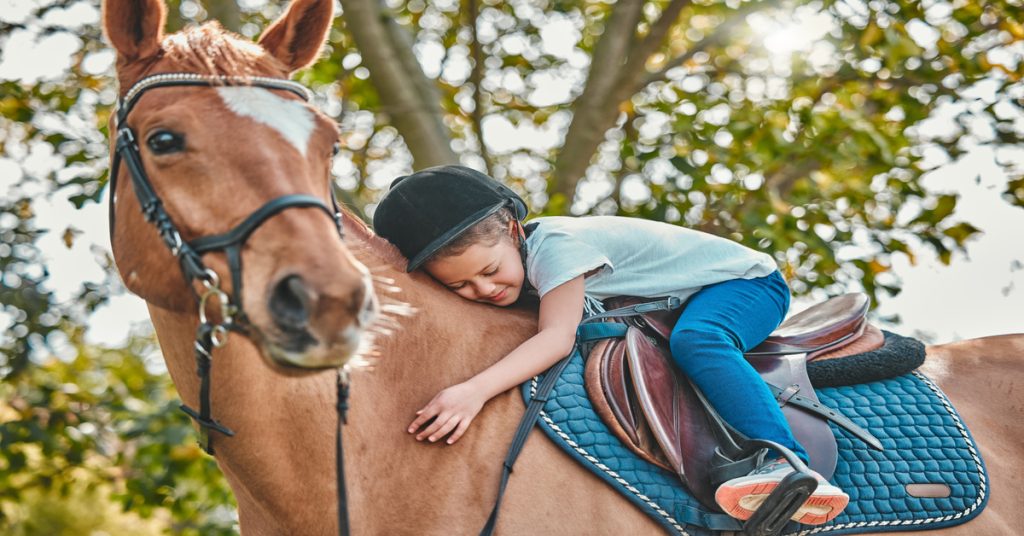 A young girl wearing a helmet sitting on the saddle of a chestnut horse, leaning forward to give it a hug.