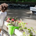 A small child standing beside container planters on a dock over a lake and watching a white boat move through the water.