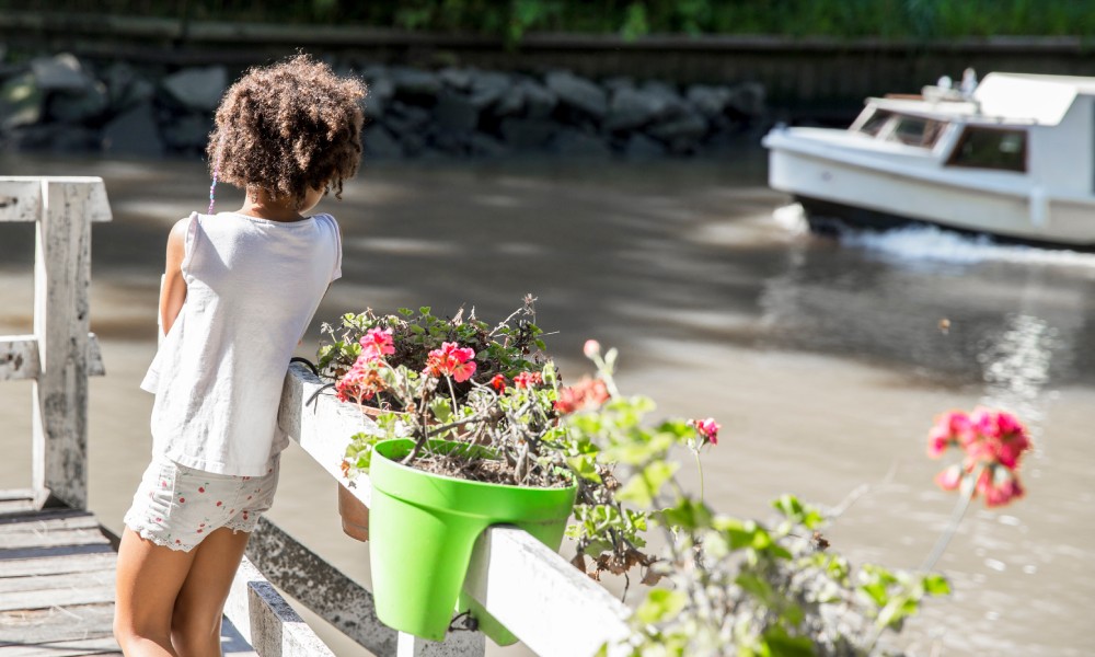 A small child standing beside container planters on a dock over a lake and watching a white boat move through the water.