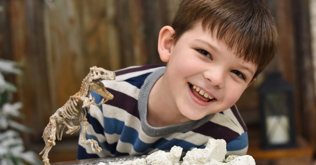 A smiling young boy in a striped shirt standing in front of a table with a model dinosaur and chalky clay on it.