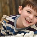 A smiling young boy in a striped shirt standing in front of a table with a model dinosaur and chalky clay on it.