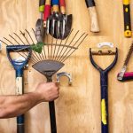 A person is grabbing a rake that is hanging on a wooden wall of an organized shed. Other tools appear around the rake.