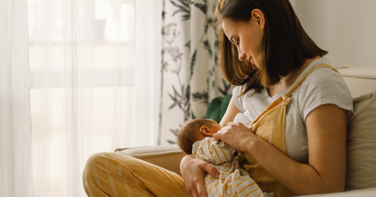 A young mom wearing a white shirt and yellow overalls sits in a cozy chair breastfeeding her newborn.