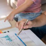 A person using a pen to reference a data sheet while their little kid sitting next to them points to a calculator.