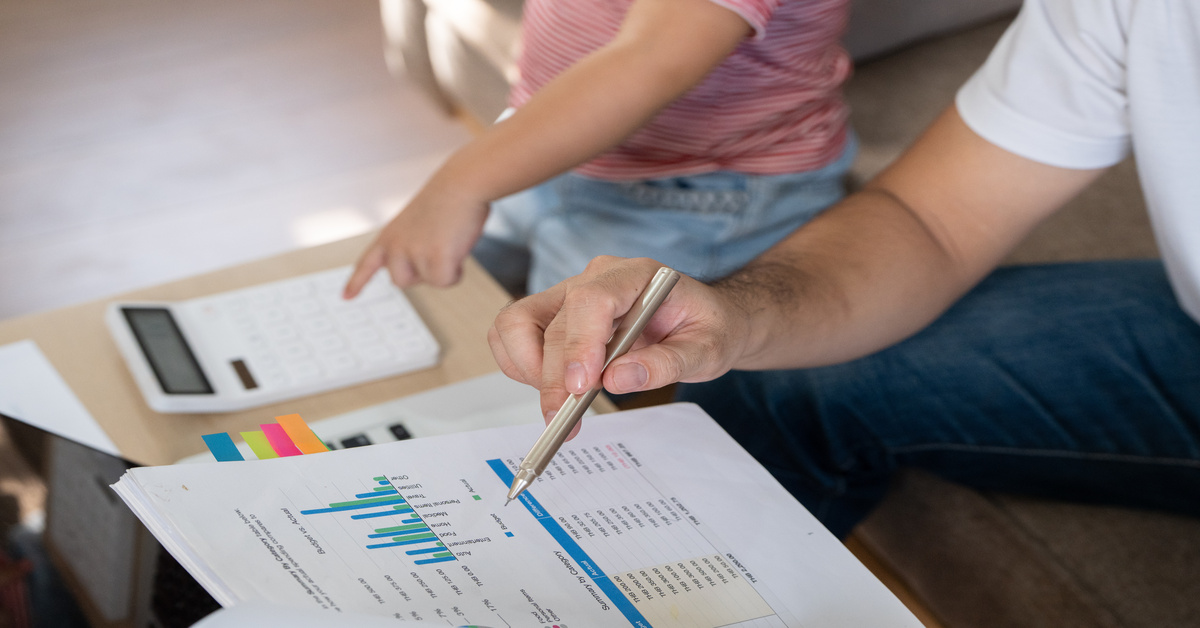 A person using a pen to reference a data sheet while their little kid sitting next to them points to a calculator.