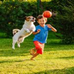 A boy runs on grass as a small dog leaps beside him toward a ball in the air. Trees and shrubs are in the background.