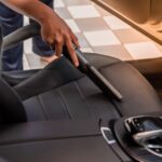 A close-up of a person's hand holding a vacuum cleaner tube extension to clean the interior seats of a car.