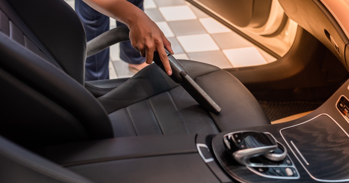 A close-up of a person's hand holding a vacuum cleaner tube extension to clean the interior seats of a car.