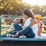 A mother and daughter sit on a colorful playground merry-go-round, laughing together in warm afternoon light.