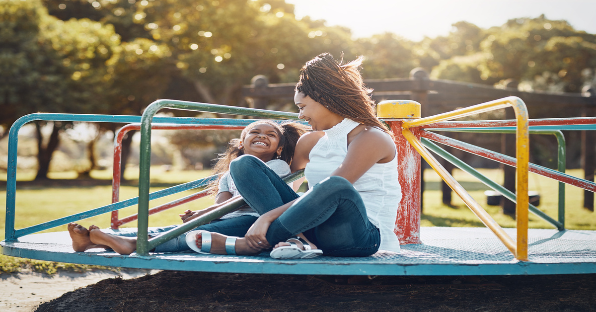 A mother and daughter sit on a colorful playground merry-go-round, laughing together in warm afternoon light.