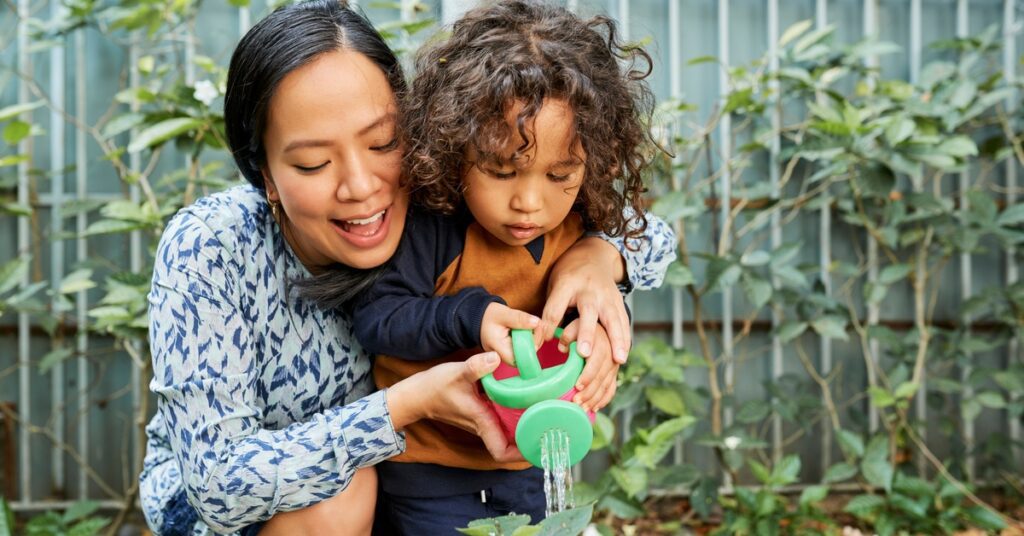 A close-up of a woman and a small kid, holding a green plastic container to water a small plant on the ground.