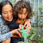 A close-up of a woman and a small kid, holding a green plastic container to water a small plant on the ground.