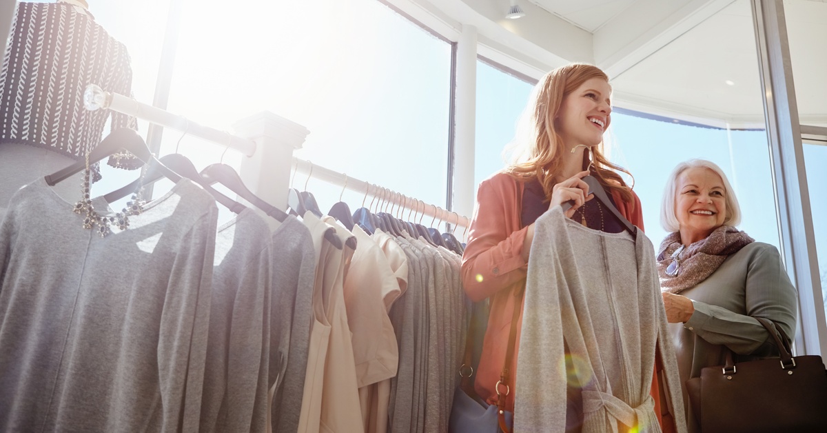 Young and older woman browse dresses together in bright clothing store, sunlight streaming through large windows.