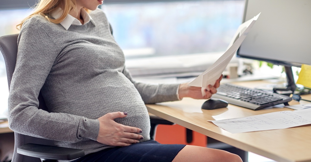 A pregnant woman at a desk in an office. With one hand, she holds a piece of paper, with the other, she cradles her belly.